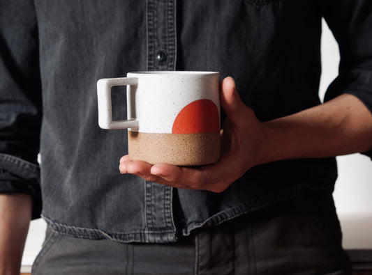 A handmade ceramic mug with an orange half circle pattern and a square handle