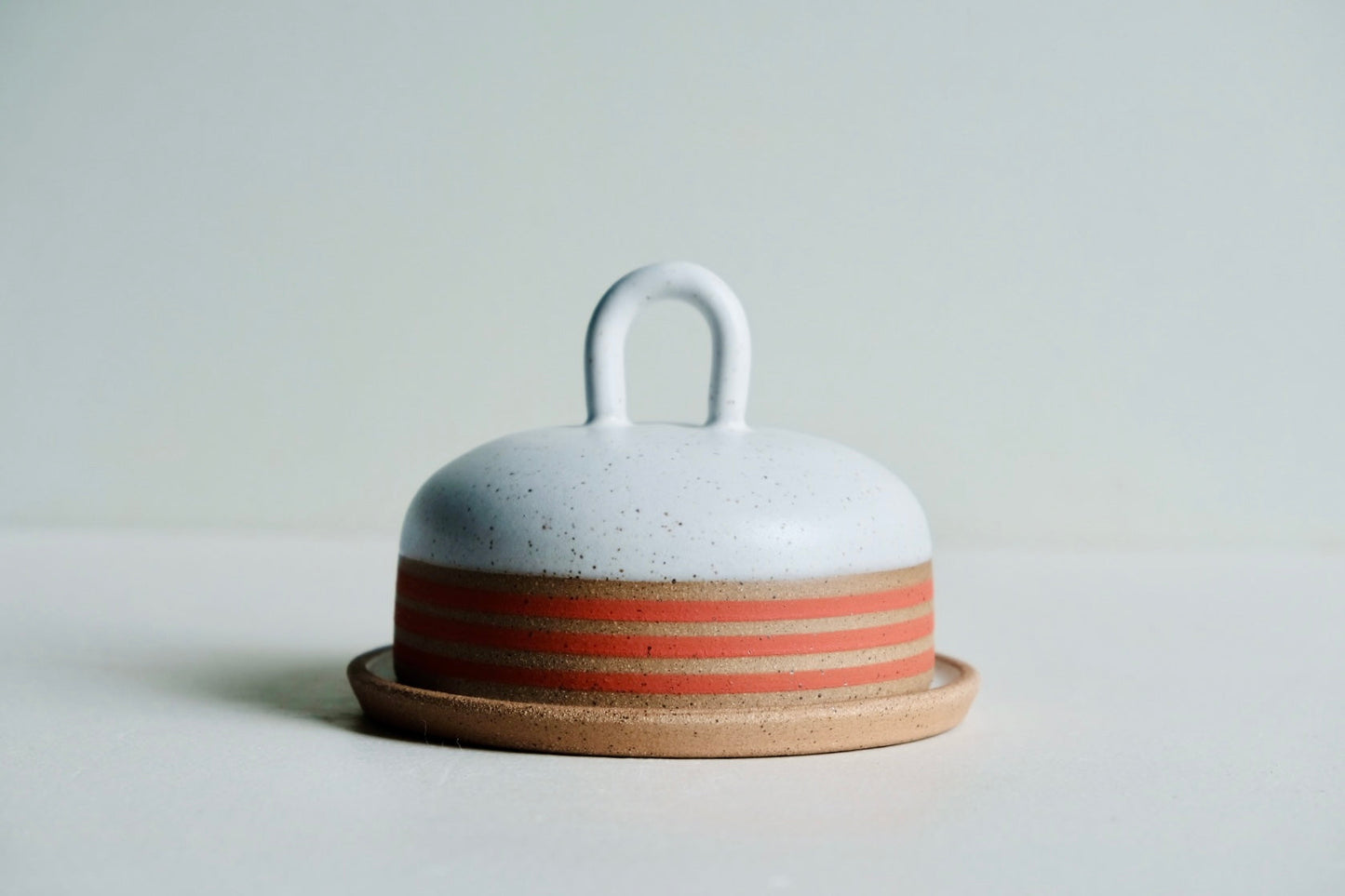 A round butter dish dipped in white satin glaze with a ring of red stripes at the bottom. A cute little handle, too.