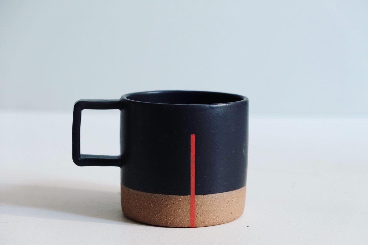 A handmade ceramic mug with a square handle, fully dipped in black with a simple red line.