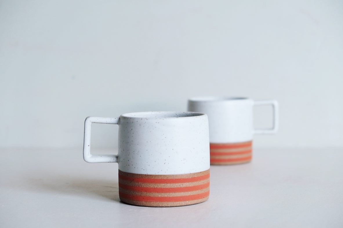 A handmade ceramic coffee mug with a square handle and linear, poppy-colored design.