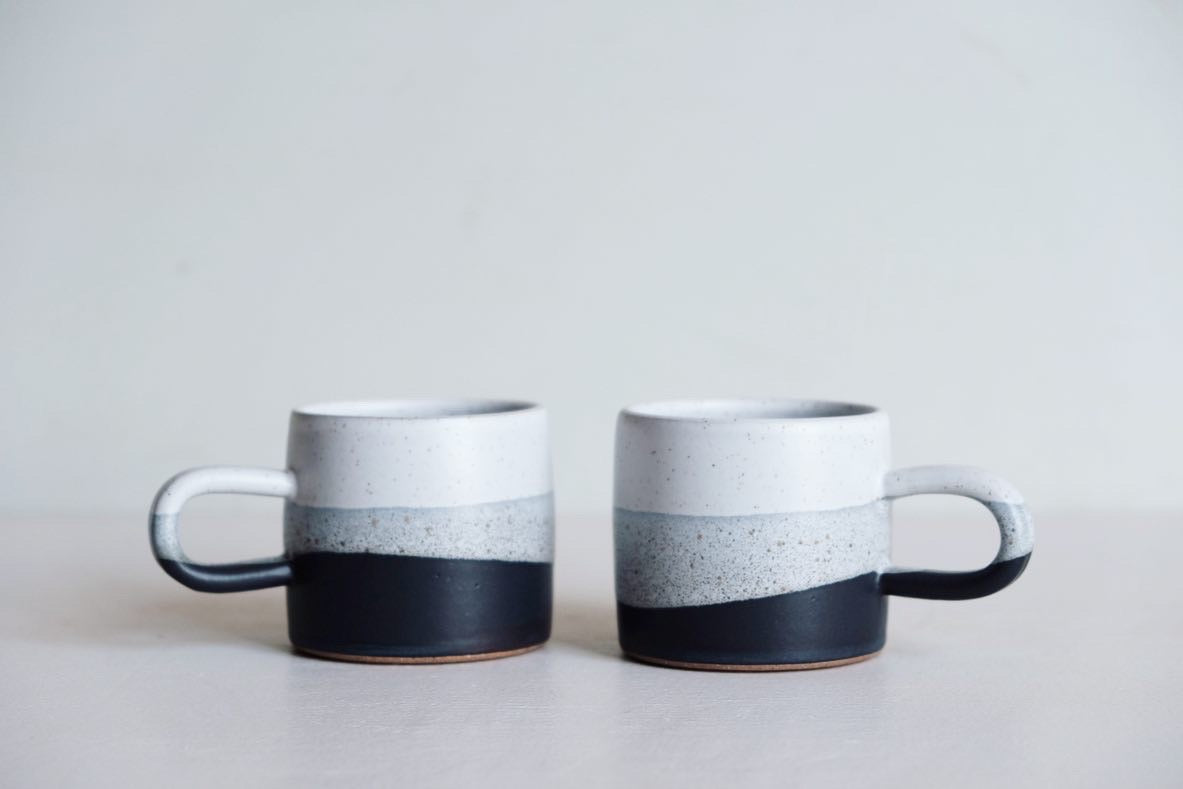 A little mug with a round handle dipped in black, grey and white speckled glaze reminsicent of wet sand on a beach